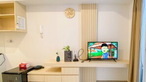 A modern living room with a flat-screen TV, a small desk, a clock, and a plant. - Good And Cozy Studio Apartment At Tokyo Riverside Pik 2 (Teluknaga)