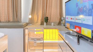 Une chambre moderne dotée d’un grand téléviseur à écran plat affichant diverses icônes d’applications, d’une chaise blanche, d’une petite table et d’une fenêtre avec des rideaux.