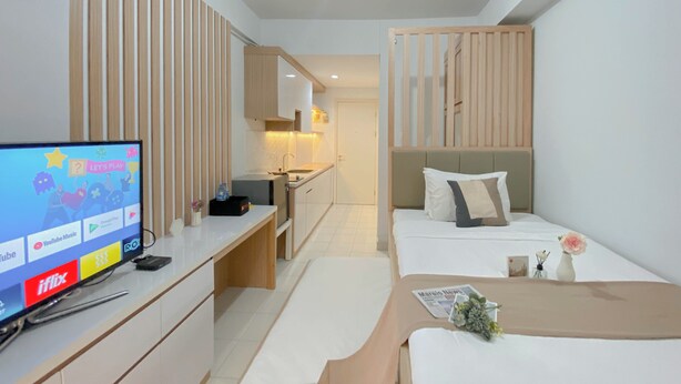 Une chambre d’hôtel moderne équipée d’une télévision diffusant une chaîne pour enfants, d’une kitchenette, d’un lit recouvert d’une courtepointe beige et d’une petite table agrémentée d’un vase de fleurs.