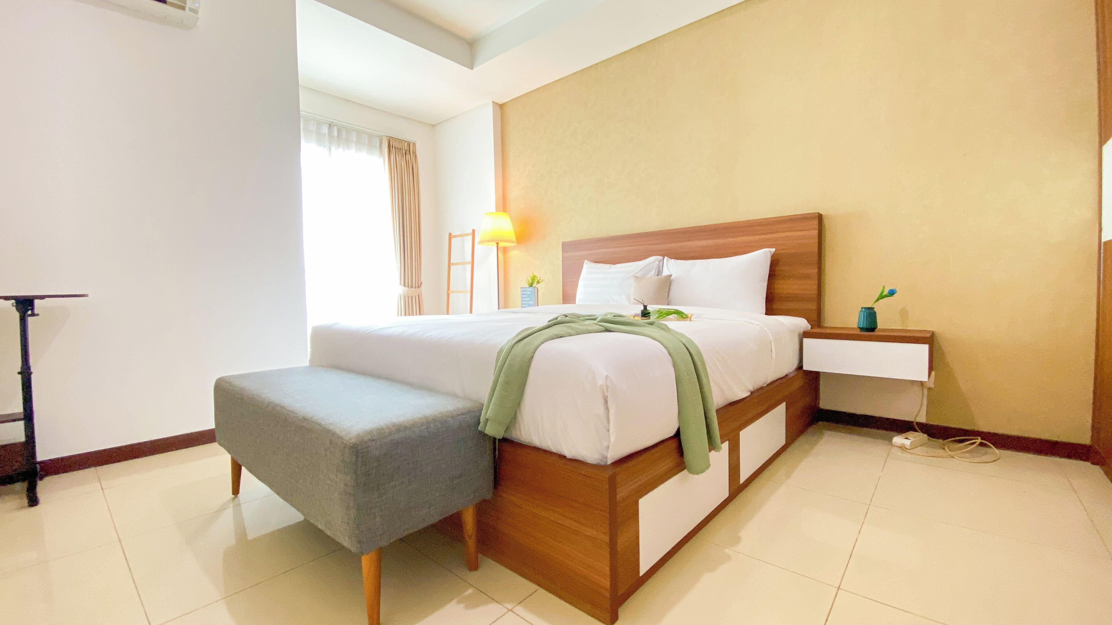 A hotel room with a bed, bedside table, lamp, and a small stool.