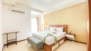 A hotel room with a bed, bedside table, bench, lamp, and an air conditioning unit. - Homey And Comfort 2Br Green Bay Condominium Apartment (Jakarta)
