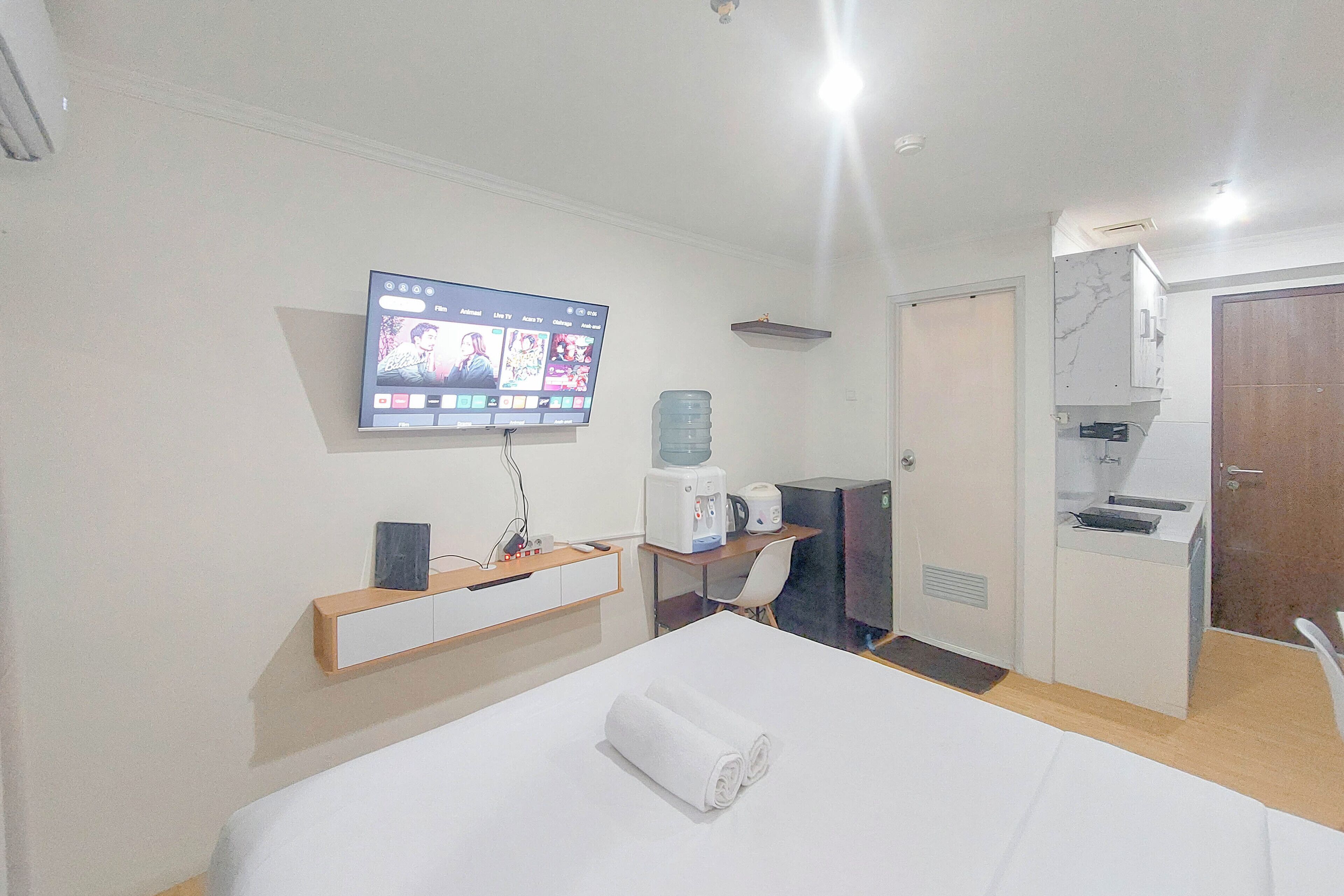 Habitación de hotel con cama, televisión montada en la pared, escritorio con silla, dispensador de agua y una kitchenette pequeña.