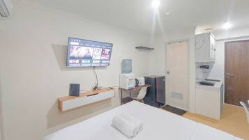 Habitación de hotel con cama, televisión montada en la pared, escritorio con silla, dispensador de agua y una kitchenette pequeña.