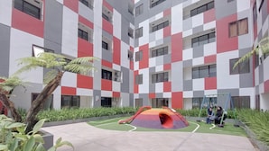 Un edificio de varios pisos con fachada a cuadros, una zona de juegos infantiles con un tobogán y un pequeño jardín con palmeras.