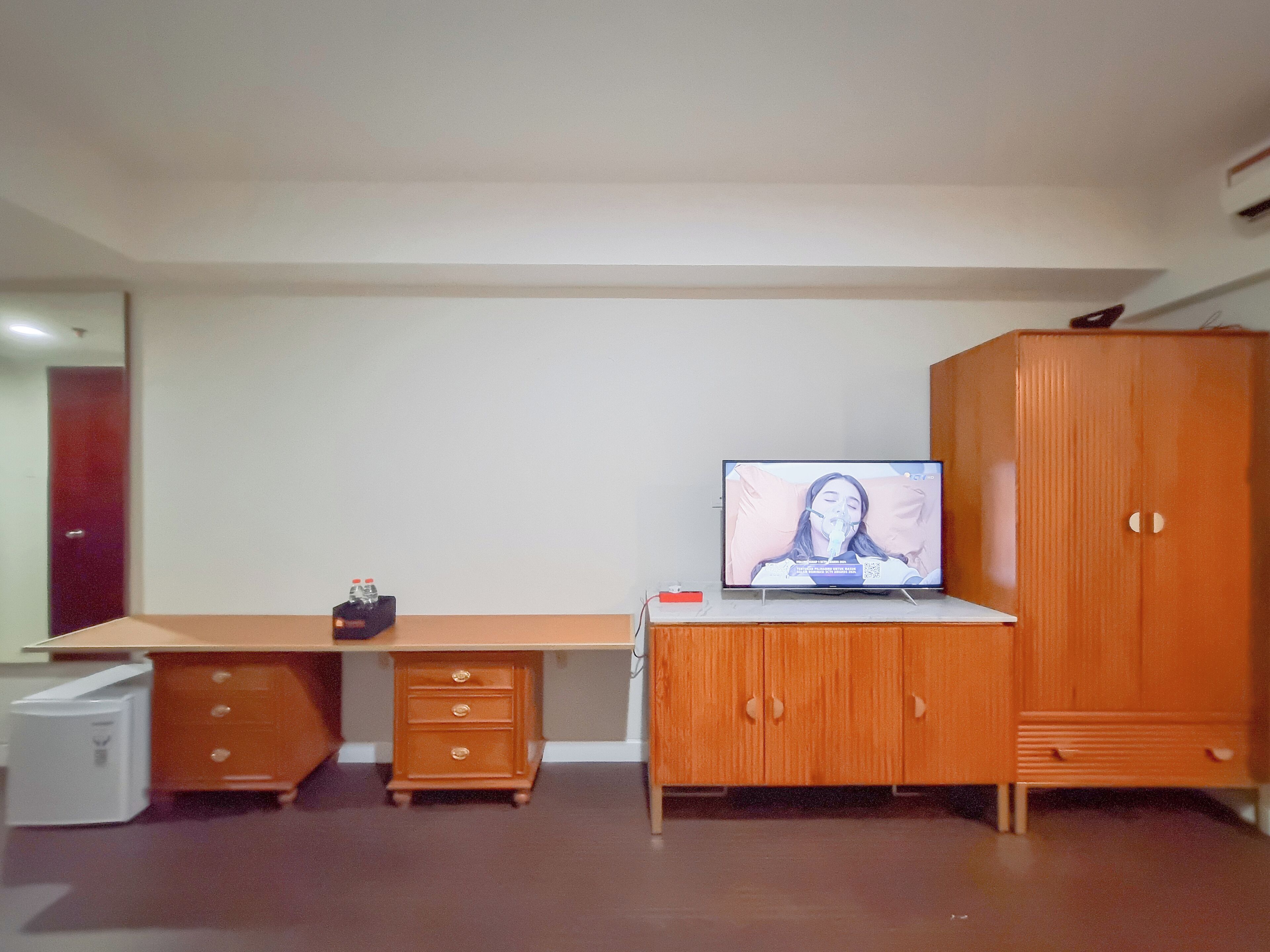 A room with a wooden desk, a television, a wardrobe, and a small white appliance.