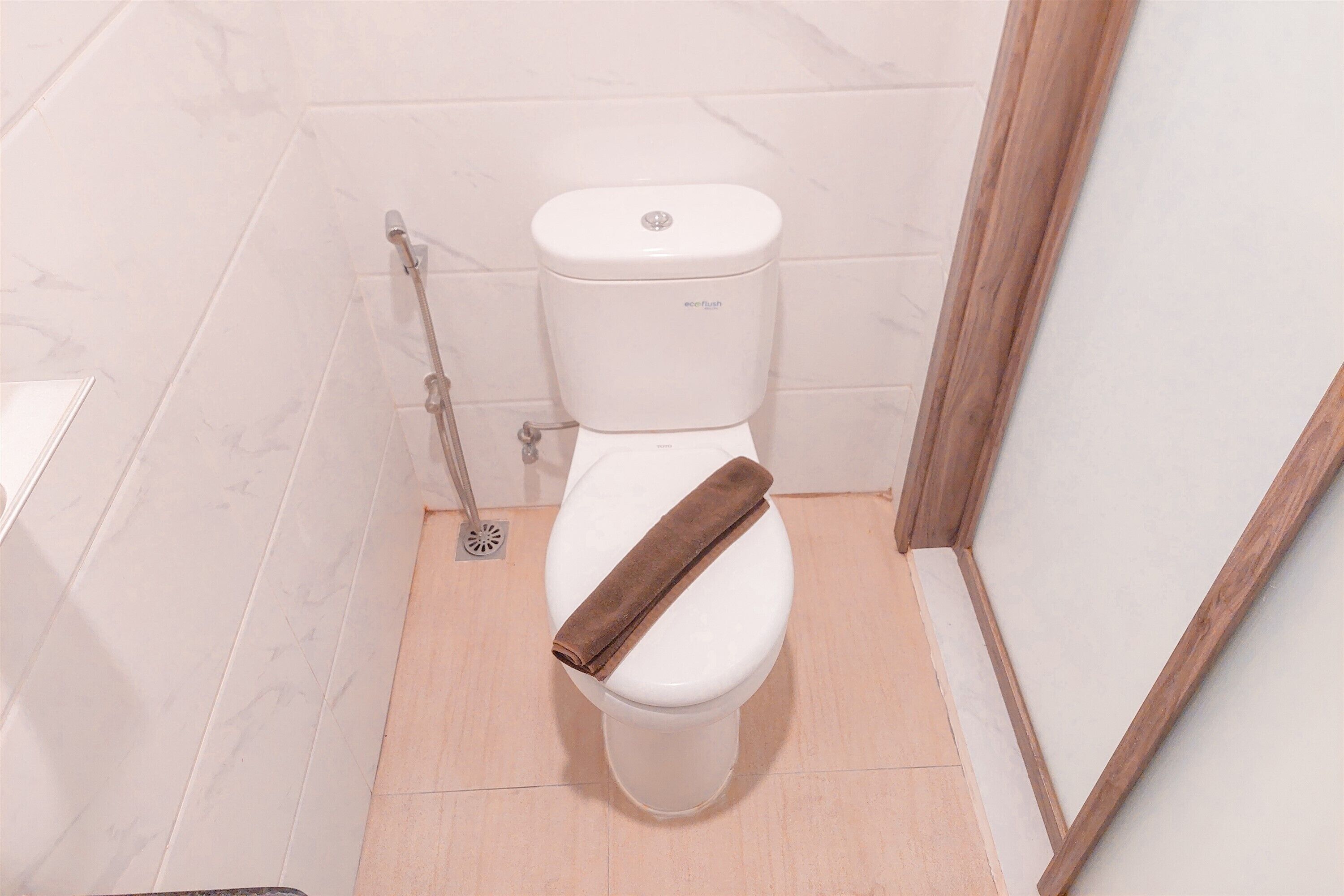 A white toilet with a brown towel on the seat, a plunger to the left, and a wooden frame leaning against the wall.