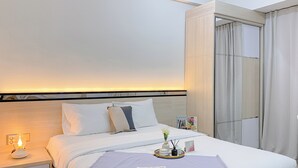 Una cama bien hecha con ropa blanca, una mesita de noche con una lámpara y una bandeja con un florero y un marco de fotos.