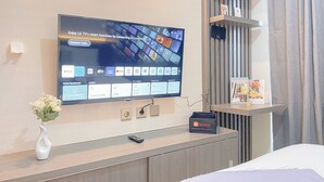 A modern hotel room with a large flat-screen TV mounted on the wall, a remote control on the console, and a vase with flowers on the console. - Cozy Living Studio At Vasanta Apartment (Cikarang)