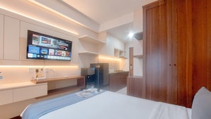 A modern hotel room with a large bed, a flat-screen TV mounted on the wall, a wooden wardrobe, and a small desk with a chair. - Minimalist And Warm Studio Apartment At B Residence (Pagedangan)