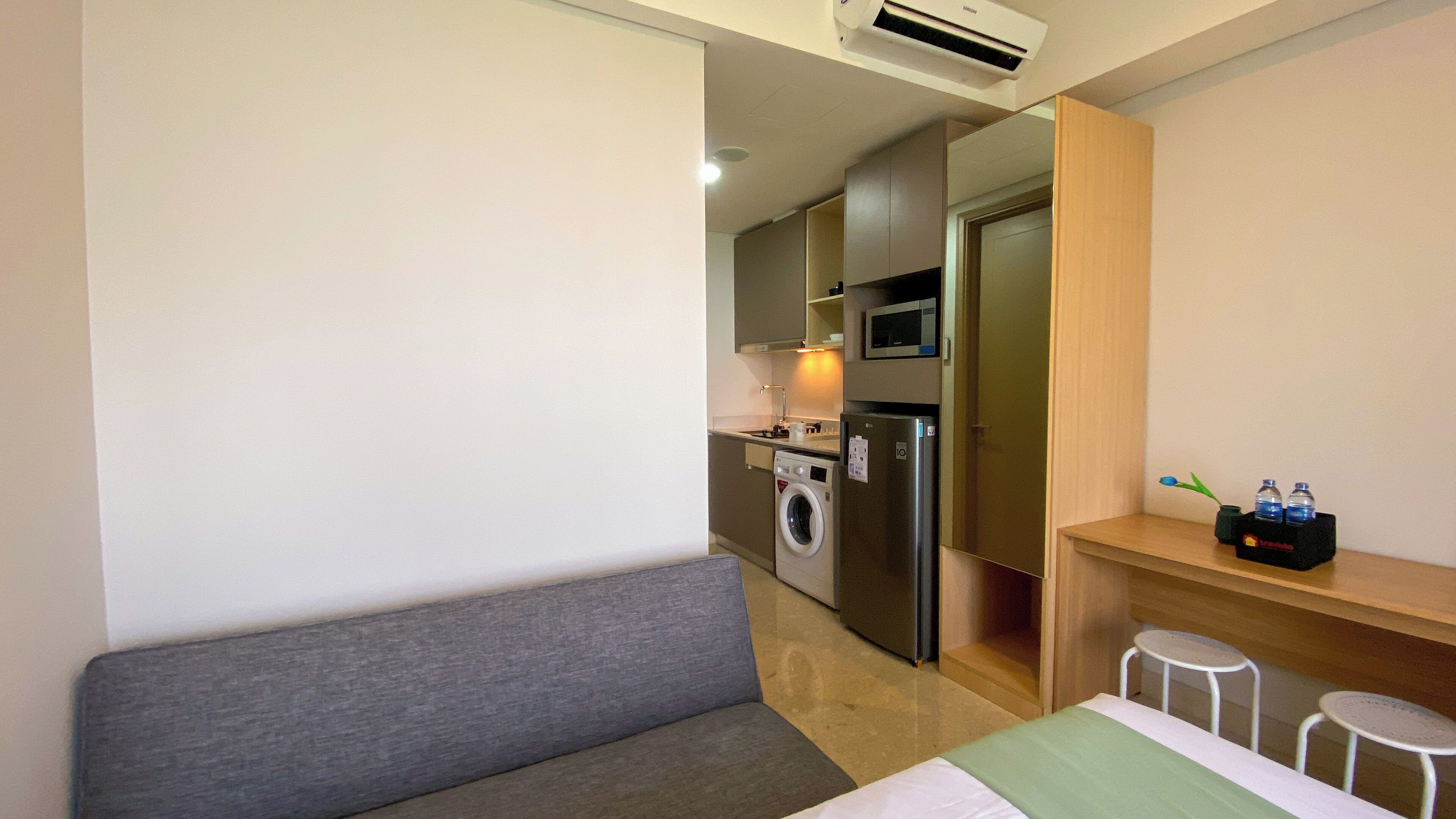 A compact hotel room with a bed, a small seating area, a kitchenette with a microwave and a washing machine, and a shelf with water bottles.