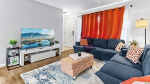 Un salon moderne avec un téléviseur à écran plat, un canapé bleu, un pouf à motifs et une grande fenêtre ornée de rideaux orange.