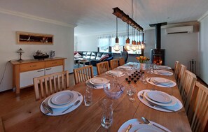 Une salle à manger avec une table en bois dressée pour six personnes, entourée de chaises. On aperçoit une cheminée au fond de la pièce, et un tableau est accroché au mur.
