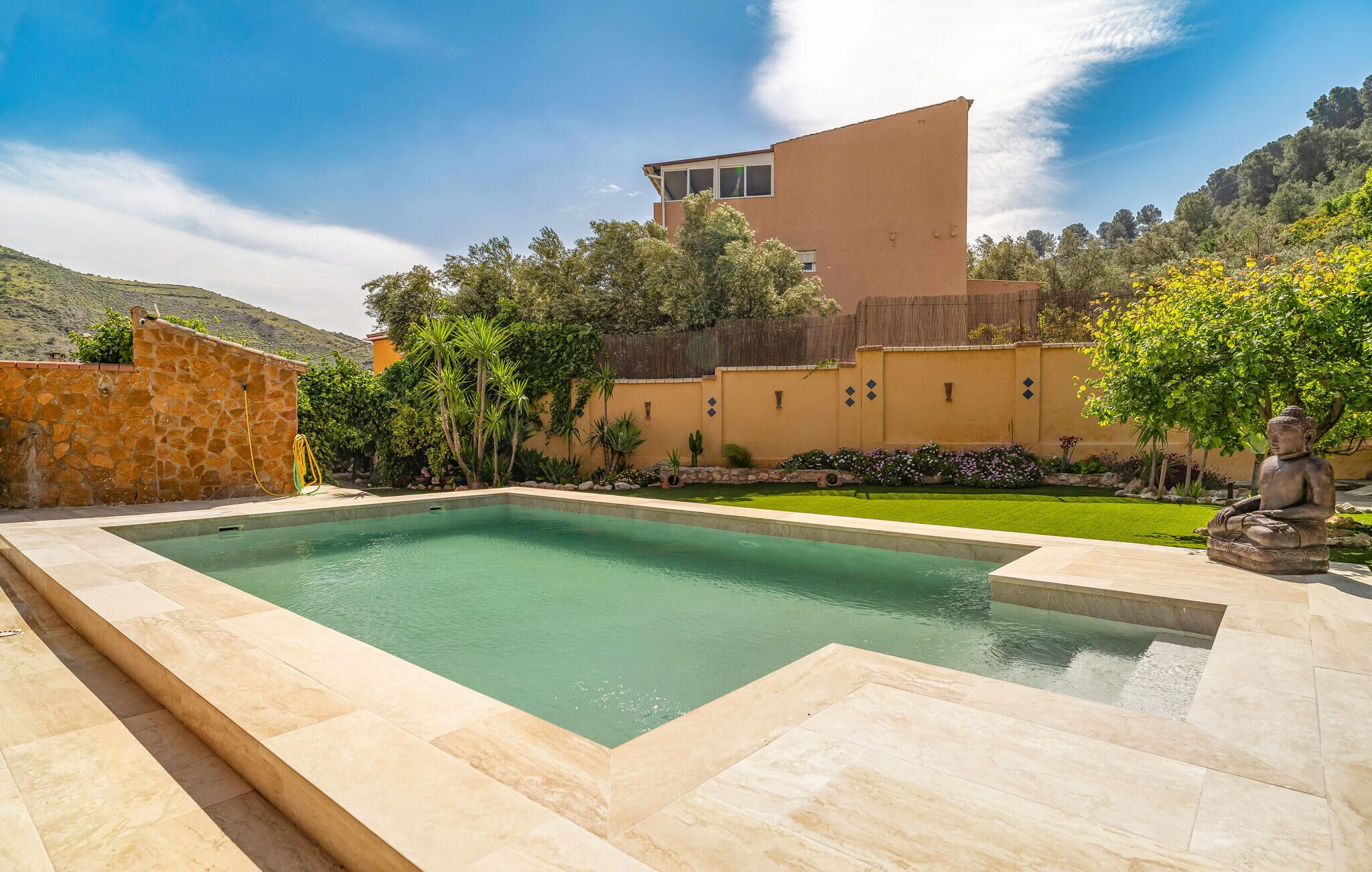 A rectangular swimming pool with a stone border, surrounded by a well-maintained garden with various plants and a Buddha statue.