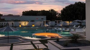 Zona de piscina amb gandules i ombrelles, envoltada d'un jardí paisatgístic i un edifici al fons.
