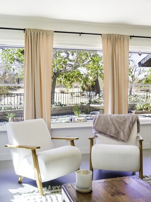 A room with two white armchairs, a wooden table, and a view of a garden through large windows with beige curtains. - Modern Desert Retreat on 5 Acres with Fire Pit! (Yucca Valley)