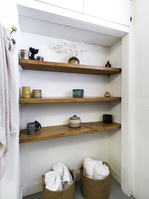 A corner with three wooden shelves, each holding various items like candles, a vase, and containers. There are two brown baskets at the bottom with towels.