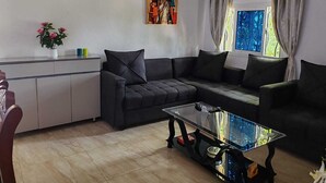 Een woonkamer met een bank, een glazen salontafel, een schilderij aan de muur en een raam met gordijnen.