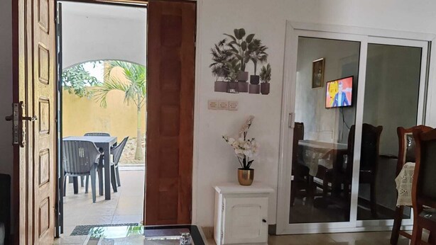 Een open deur die leidt naar een terras met een tafel en stoelen, een klein wit kastje met een bloempot, en een televisie op een schuifdeur van glas.