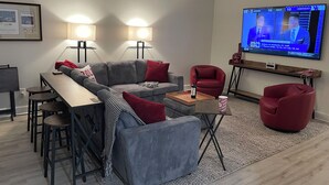A modern living room with a sectional sofa, a dining table with bar stools, a red armchair, and a flat-screen TV displaying a sports game.