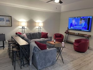 Una sala de estar moderna con un sofá modular, una mesa de comedor con taburetes de bar, un sillón rojo y un televisor de pantalla plana mostrando un partido deportivo.