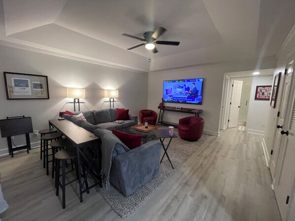A modern living room with a grey sectional sofa, red armchairs, a dining area with a wooden table and chairs, a flat-screen TV mounted on the wall, and a ceiling fan. - New remodeled 2BR 2BT condo, only 1.4 miles to Bryant-Denny Stadium! ROLL TIDE!! (Tuscaloosa)