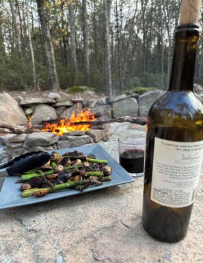 Une bouteille de vin, une assiette de légumes grillés et un verre de vin rouge, le tout posé sur une surface en pierre, avec un feu en arrière-plan.