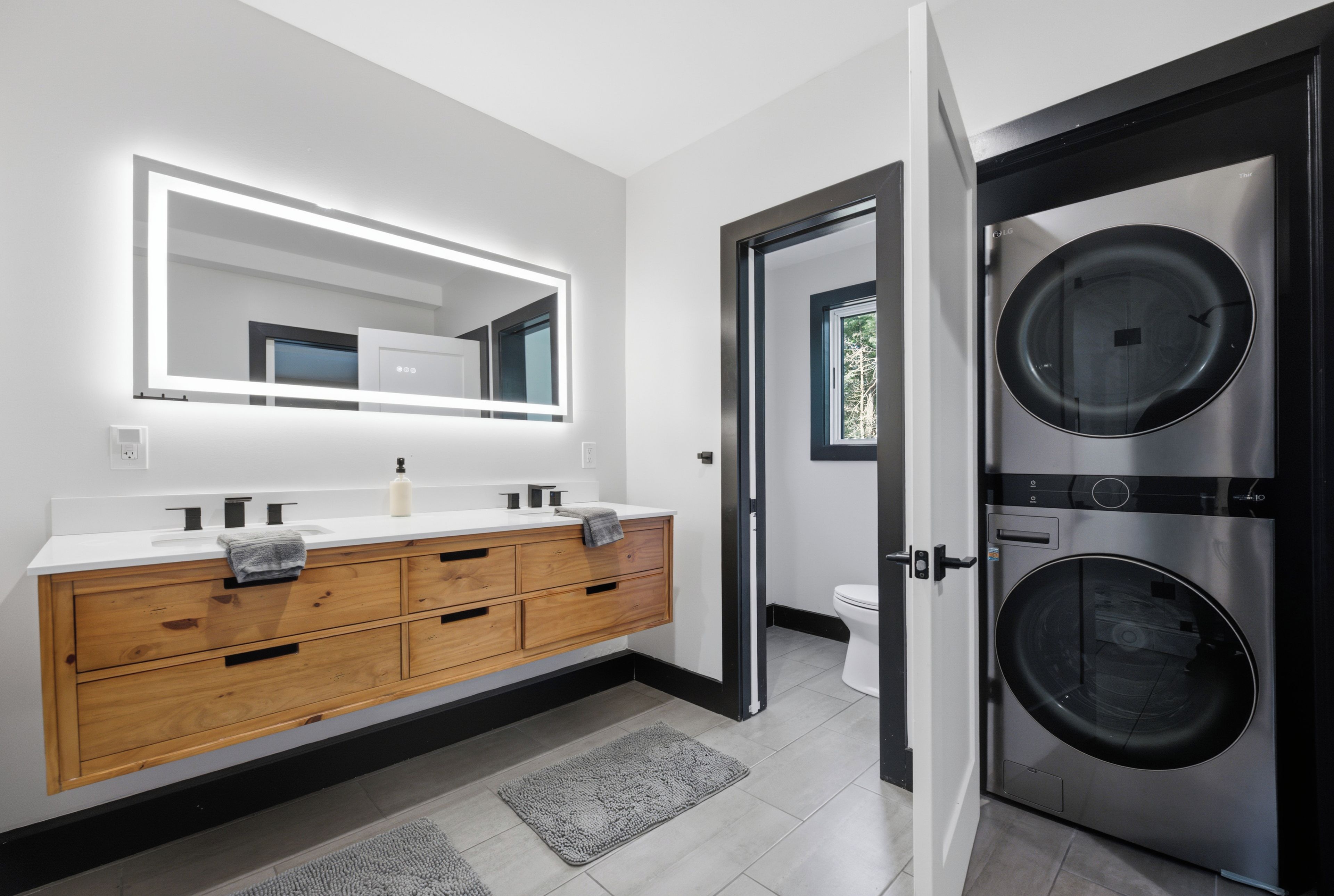 A modern bathroom with a wooden vanity, a large mirror, a toilet, and a double-door laundry machine.