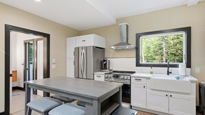 Een moderne keuken met een koelkast, oven en kookplaat van roestvrij staal, een raam met uitzicht op bomen en een eethoek met een tafel en twee stoelen.