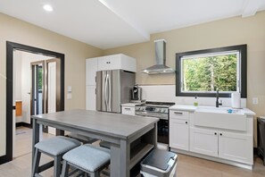 Una cucina moderna con frigorifero, forno e piano cottura in acciaio inossidabile, una finestra con vista sugli alberi e un'area pranzo con tavolo e due sedie.