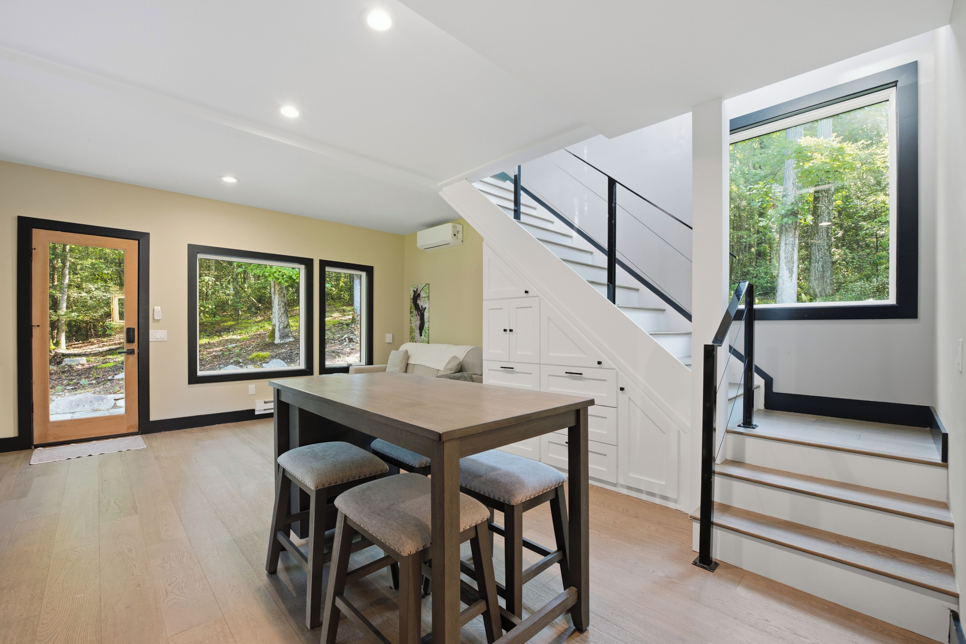 A modern kitchen with a wooden table and bar stools, a staircase with a glass railing, and large windows with forest views.
