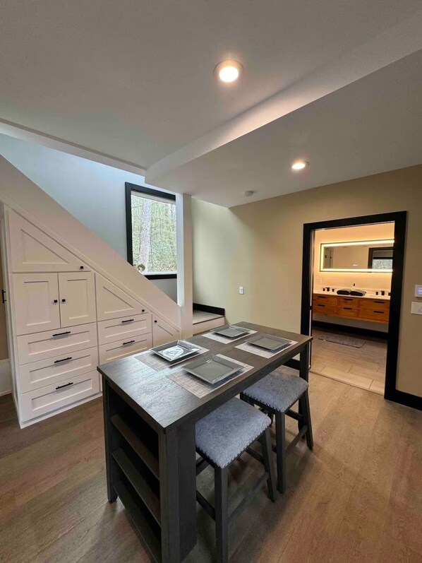 Un coin repas avec une table et des chaises en bois foncé, un escalier et une cuisine visible à travers une porte ouverte.