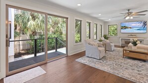 A spacious living room with a large sliding glass door leading to a balcony, a comfortable seating area with a sofa and armchairs, a flat-screen TV mounted on the wall, and a ceiling fan.