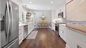 A modern kitchen with white cabinetry, a stainless steel refrigerator, a dining area with a chandelier, and a large painting on the wall.