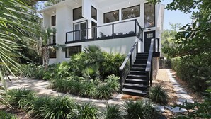 A modern two-story house with a balcony, surrounded by lush greenery and a stone pathway.