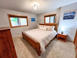 A bedroom with a wooden bed, two windows, a nightstand with a lamp, and a picture on the wall.