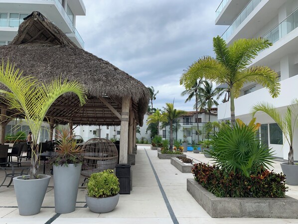 A modern hotel courtyard with a thatched-roof gazebo, potted plants, outdoor seating, and a pool area.