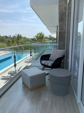 Un balcon moderne doté d’une balustrade en verre, donnant sur une piscine, aménagé avec un fauteuil et un pouf confortables, et offrant une vue sur une végétation luxuriante.