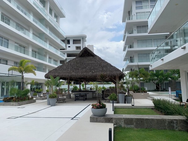 Une cour d’hôtel moderne, dotée d’une structure centrale à toit de chaume, entourée de plantes en pot et de mobilier d’extérieur.