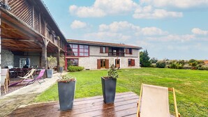 Una terraza de madera con mobiliario exterior y plantas en macetas, un edificio de piedra con contraventanas rojas y un césped.