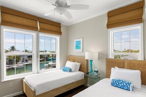 Une chambre d’hôtel avec deux lits, un ventilateur de plafond, de grandes fenêtres donnant sur un port de plaisance et une petite table avec une lampe.