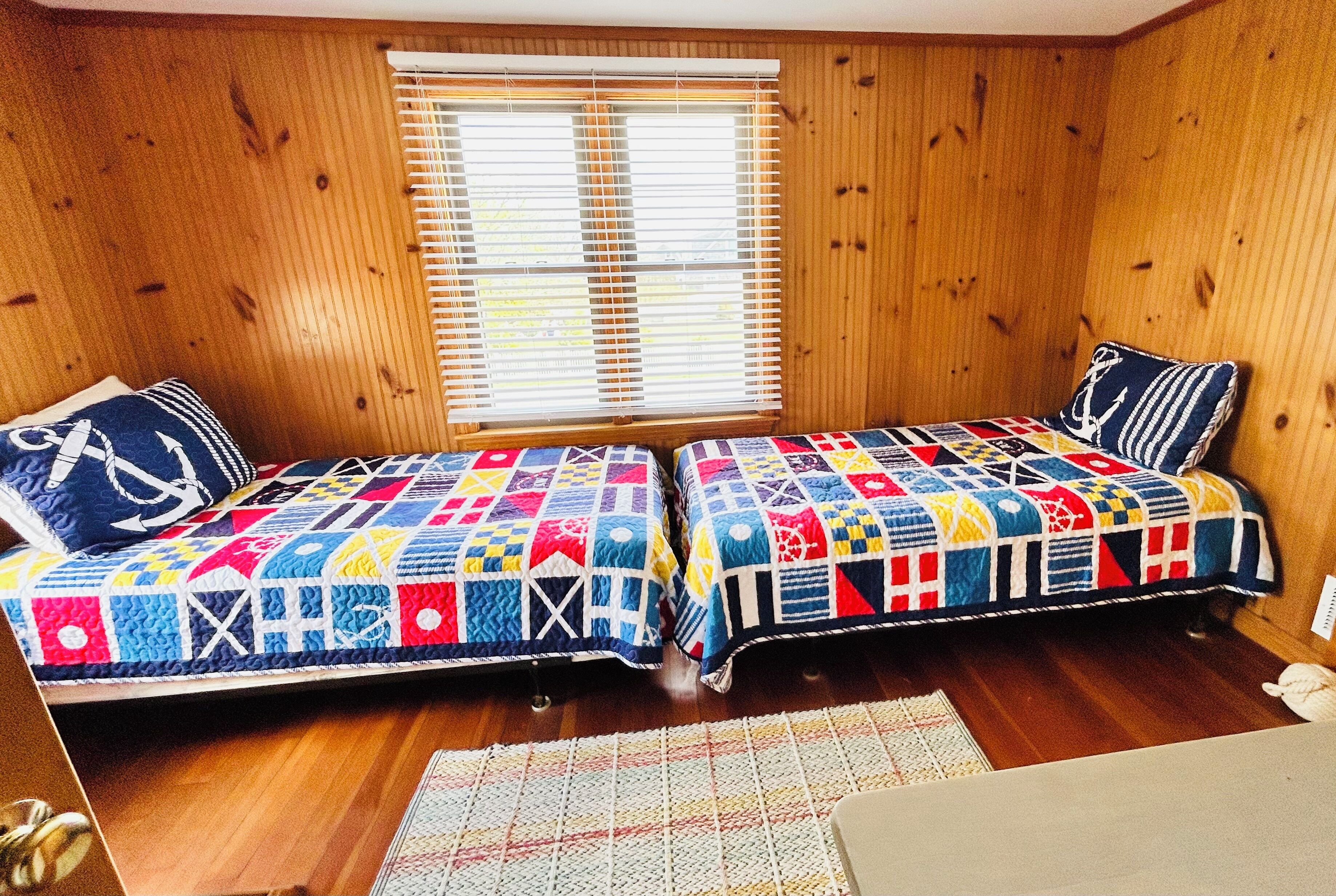 A room with two beds, wooden walls, a window with blinds, and a rug.