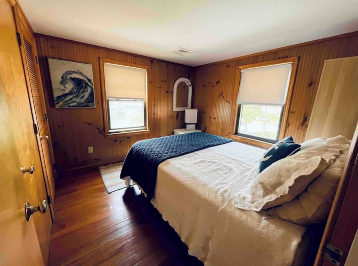 Un dormitorio con paredes de madera, una cama con ropa de cama blanca y oscura, dos ventanas con persianas, una mesita de noche con una lámpara y una puerta con pomo de latón.