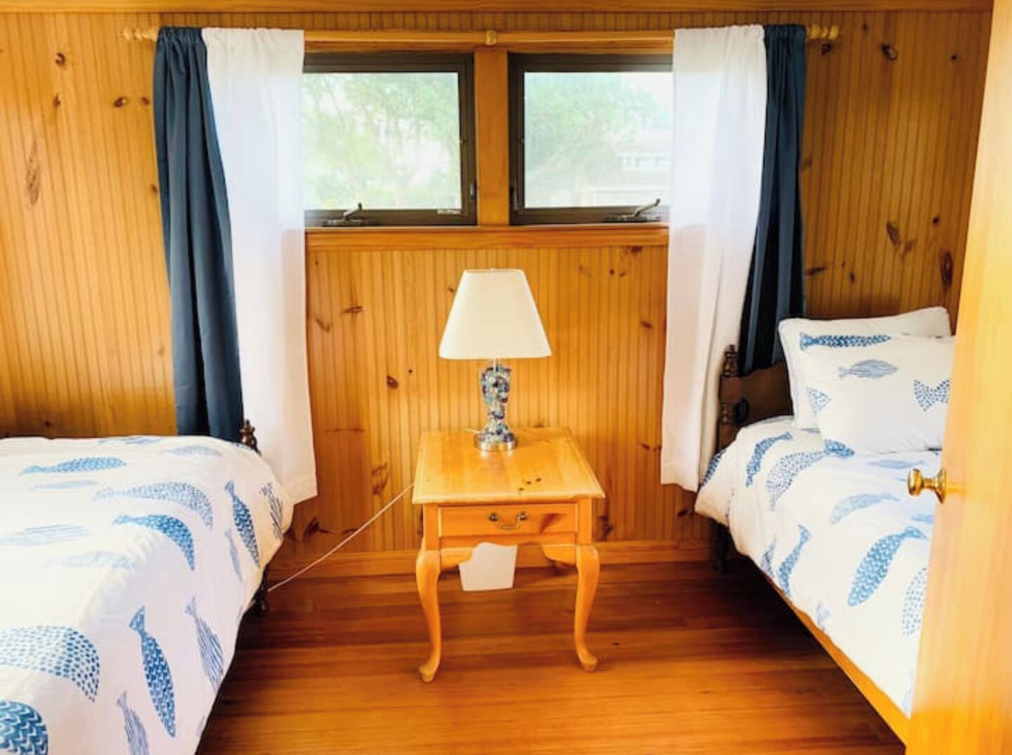 A room with two beds, a wooden nightstand with a lamp, and a window with curtains.