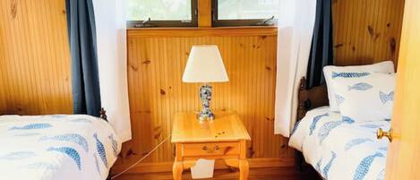A room with two beds, a wooden nightstand with a lamp, and a window with curtains.