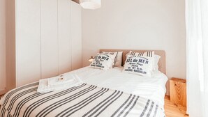 Una cama bien hecha con ropa de cama a rayas y almohadas con texto, una mesita de noche de madera y una lámpara sobre la cama.