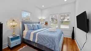 A bedroom with a bed, bedside table, lamp, TV, and windows.