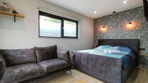 Un dormitorio moderno con un sofá gris, una cama con cubrecama azul y gris, una ventana con vistas y una lámpara de pared.