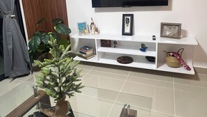 Ein Wohnzimmer mit einem an der Wand befestigten Flachbildfernseher, einem Glastisch, einem Blumentopf und einer weißen Unterhaltungseinheit mit verschiedenen Dekorationsgegenständen.