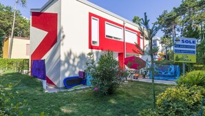 Ein modernes Gebäude mit roter und weißer Fassade, ein kleiner Gartenbereich mit einer blauen Spielanlage und ein Schild für 'Agencia Turistica Sole' mit Kontaktinformationen.