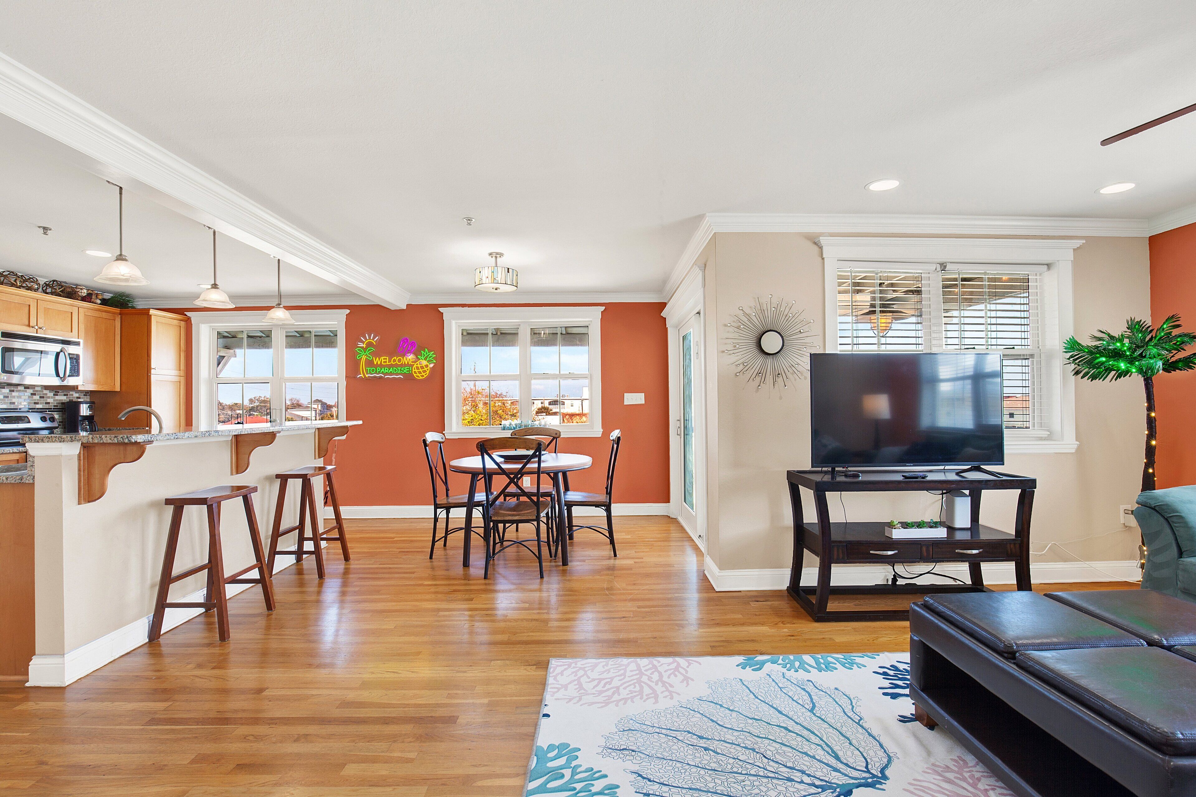A modern kitchen with wooden cabinets and a built-in microwave, a dining area with a table and chairs, a living room with a TV and a sofa, and a large window with blinds.